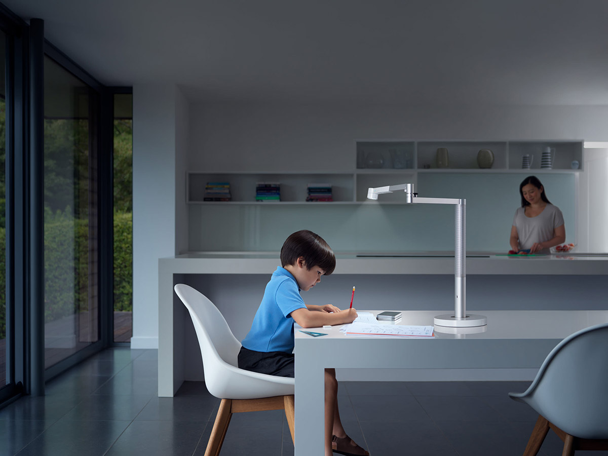 Ein Kind sitzt an einem Tisch unter einer Lampe und schreibt. Im Hintergrund steht eine Frau in einer Küche.