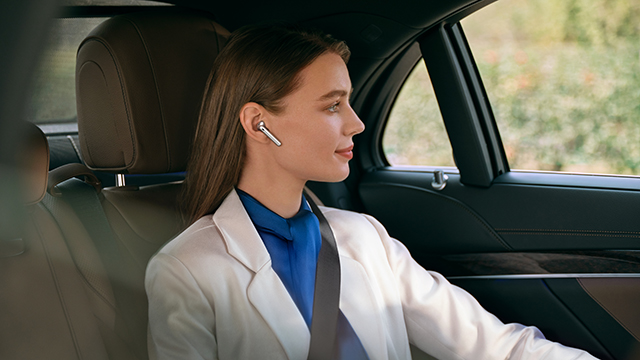 Eine Frau mit Kopfhörern sitzt im Fond eines Autos und blickt aus dem Fenster. Sie trägt eine helle Jacke und einen blauen Schal.