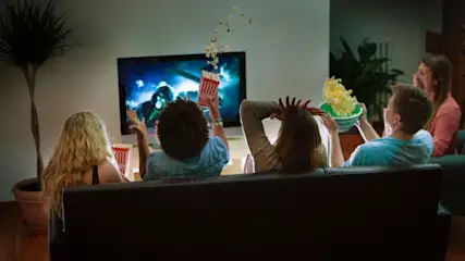 Vier Personen blicken von dunkler Couch auf Fernseher. Popcorn fliegt aus Box, andere halten Schalen. Blau beleuchtete Szene auf Bildschirm, Pflanzen im Raum.