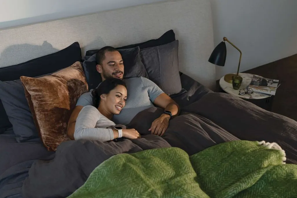 Paar liegt gemeinsam im Bett und traegt Fitnessuhren am Handgelenk