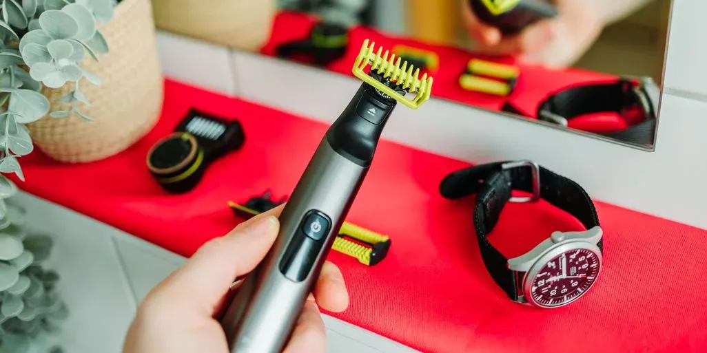 Hand hält schwarze und silberne kabellose Haarschneidemaschine mit gelb-schwarzen Aufsätzen auf rotem Tuch, daneben Uhr und Pflanze.