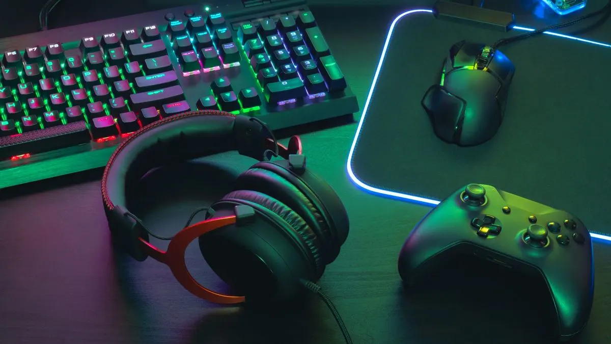 Eine beleuchtete Gaming Tastatur, ein Headset, eine Maus auf einem leuchtenden Mauspad und ein Gamecontroller liegen auf einem dunklen Schreibtisch