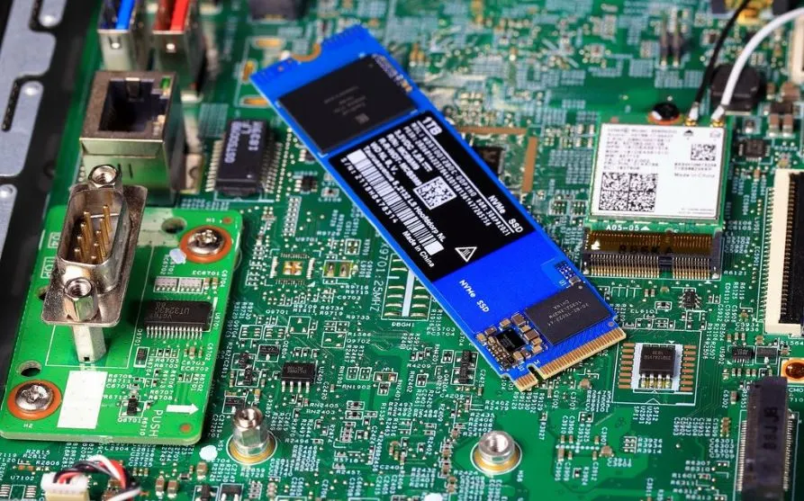 Ein blauer NVMe SSD-Stick mit 1TB-Aufschrift liegt auf einer grünen Platine mit diversen elektronischen Bauteilen und Anschlüssen.