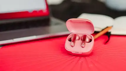 Ein offenes Ladecase mit pinkfarbenen JBL In-Ear-Kopfhörern steht auf einem roten Tisch.