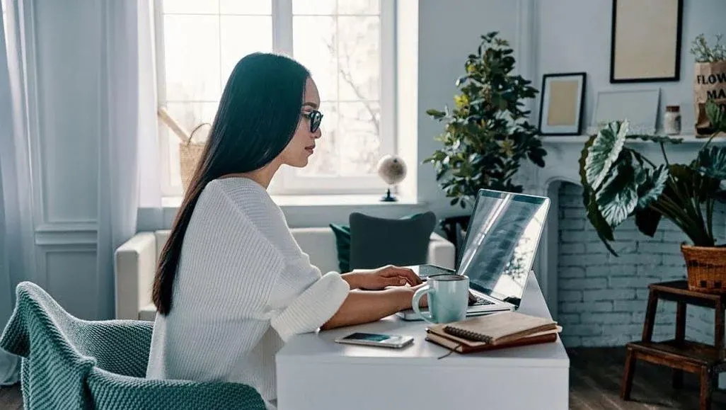 Eine Frau mit Brille tippt an einem weißen Schreibtisch auf einem Laptop. Daneben sind eine blaue Tasse, ein Smartphone und Notizbücher. Im Hintergrund ein Fenster, Pflanzen und ein Sofa.