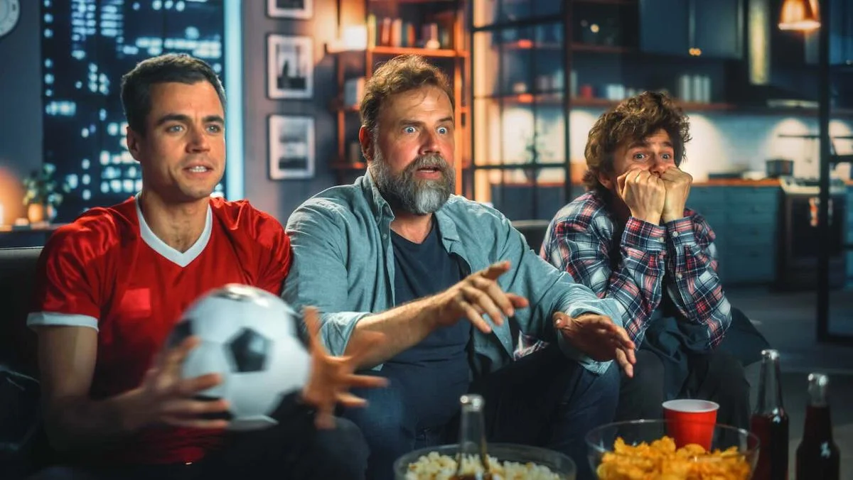 Drei Männer sitzen abends extrem angespannt auf einer Couch und schauen ein Fußballspiel. Einer hält einen Fußball in der Hand, im Vordergrund stehen Snacks und Getränke