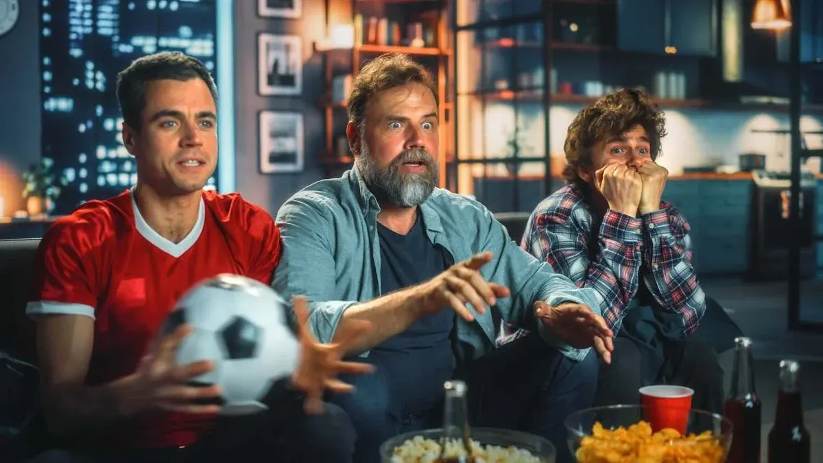 Drei Männer sitzen abends extrem angespannt auf einer Couch und schauen ein Fußballspiel. Einer hält einen Fußball in der Hand, im Vordergrund stehen Snacks und Getränke