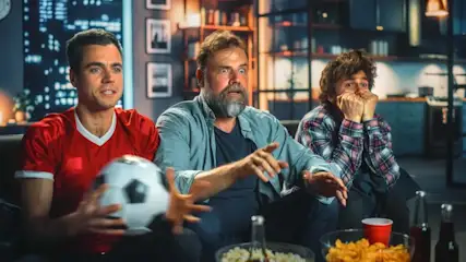 Drei Männer sitzen abends extrem angespannt auf einer Couch und schauen ein Fußballspiel. Einer hält einen Fußball in der Hand, im Vordergrund stehen Snacks und Getränke