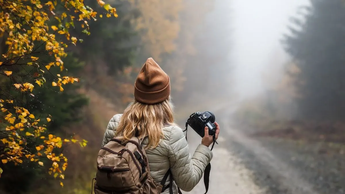 Frau mit Rucksack und Haube hält Kamera und steht auf einem nebligen Waldweg.