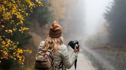 Frau mit Rucksack und Haube hält Kamera und steht auf einem nebligen Waldweg.