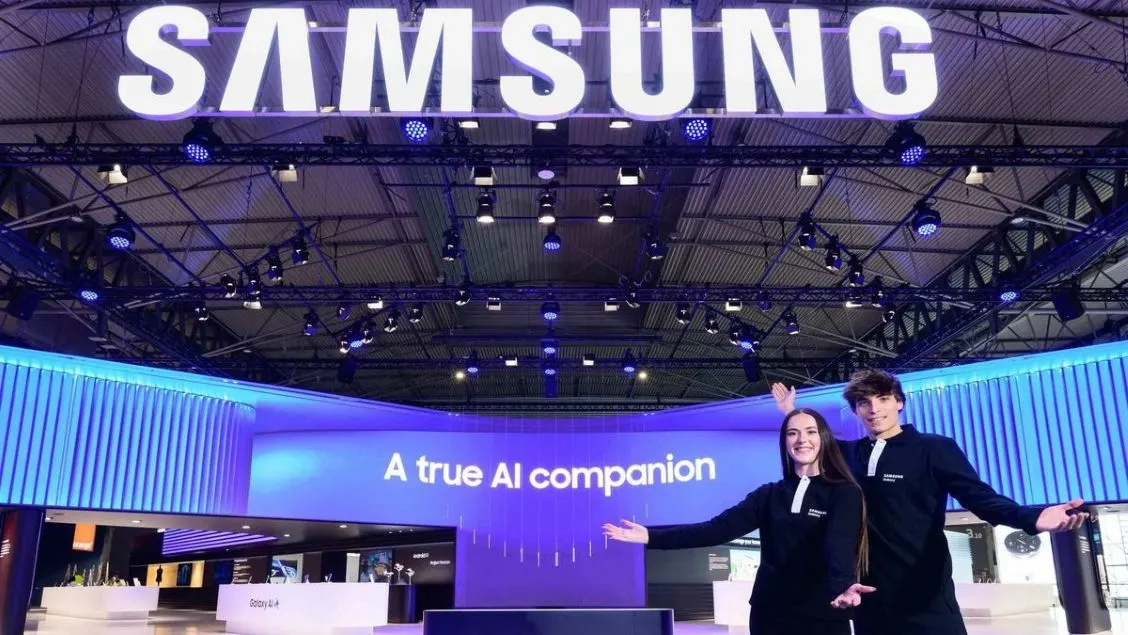 Zwei Personen posieren vor einem blauen Samsung-Stand. Ein Bildschirm zeigt "A true AI companion". Großes Samsung-Logo und Lichter oben.