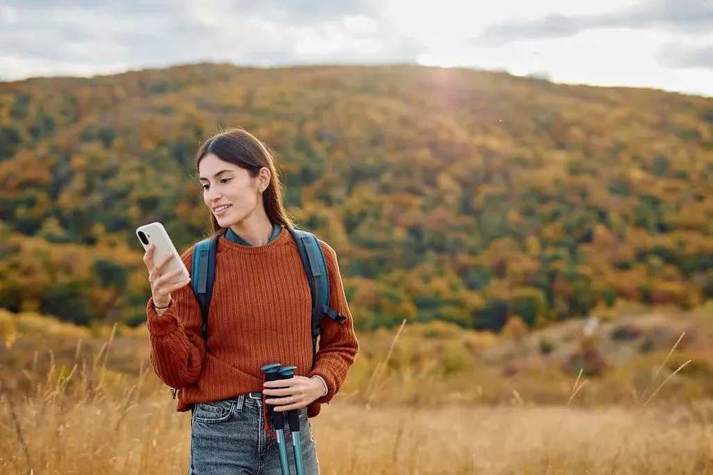 Wanderin in rotem Pullover hält Smartphone in Herbstlandschaft mit Rucksack und Fernglas.