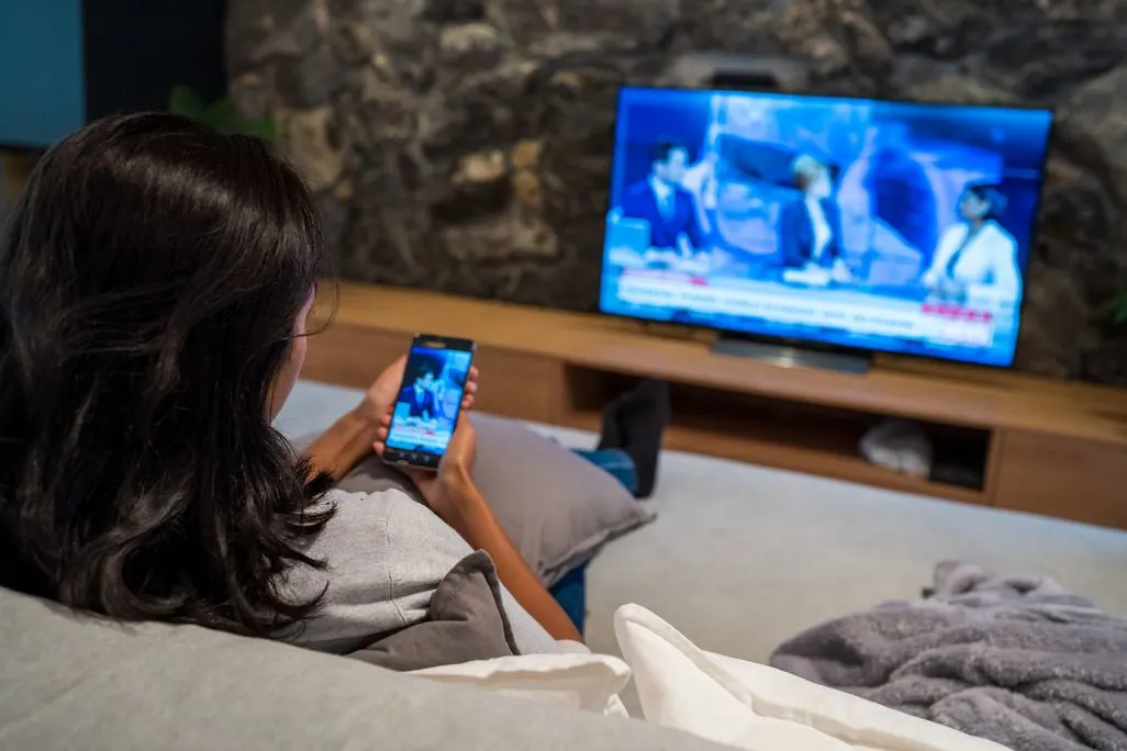Frau sitzt auf dem Sofa und schaut gleichzeitig auf ihr Smartphone und den Fernseher.