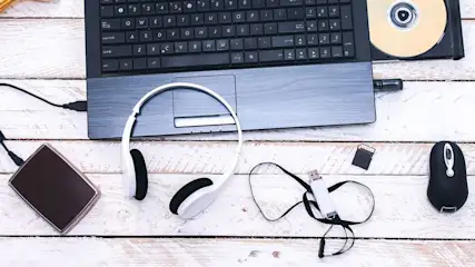 Computer-Zubehör auf Holztisch mit Laptop, Tastatur, Kopfhörern und DVD im offenen Laufwerk.