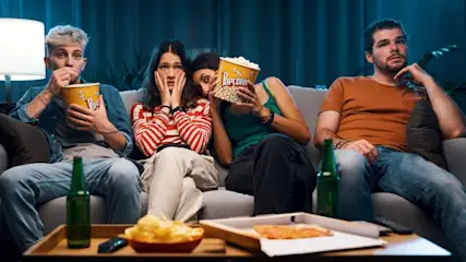 Vier Personen sitzen abends auf einem Sofa, essen Popcorn und schauen gemeinsam einen Film.
