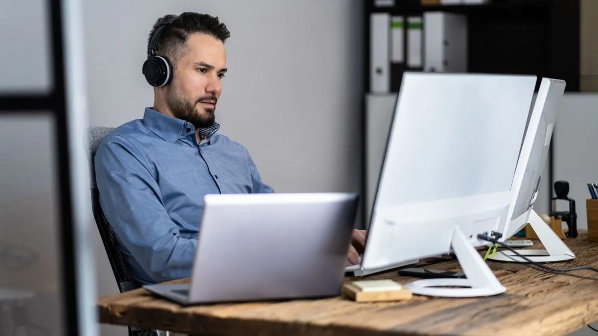 Mann im blauen Hemd mit Kopfhörern sitzt an Holztisch, arbeitet an Laptop und weißem Monitor. Im Hintergrund Ordnerregal.