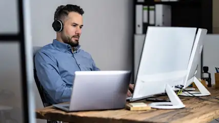Mann im blauen Hemd mit Kopfhörern sitzt an Holztisch, arbeitet an Laptop und weißem Monitor. Im Hintergrund Ordnerregal.