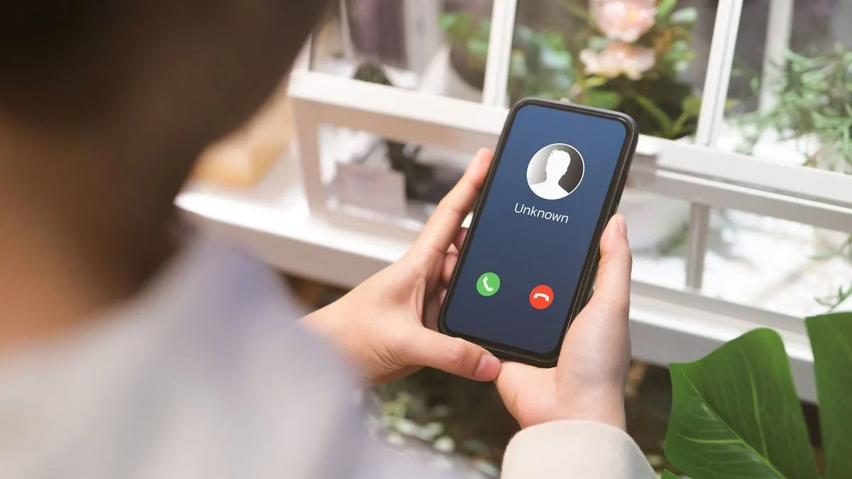 Eine Person hält ein schwarzes Smartphone, dessen Bildschirm einen unbekannten Anruf mit grünen und roten Symbolen anzeigt, vor verschwommenen Pflanzen und weißen Rahmen.