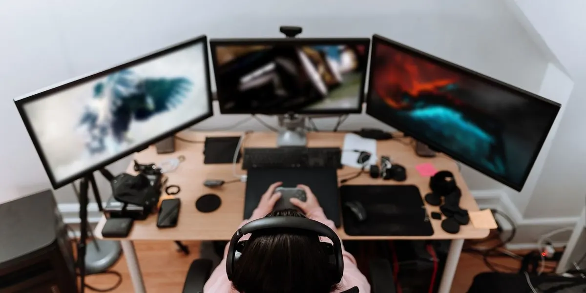 Person mit Headset sitzt an einem Schreibtisch mit drei Monitoren und spielt ein Computerspiel.