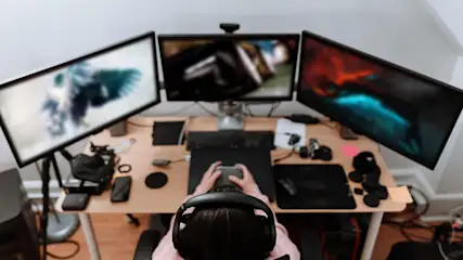 Person mit Headset sitzt an einem Schreibtisch mit drei Monitoren und spielt ein Computerspiel.