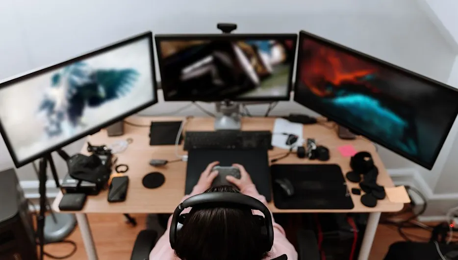 Person mit Headset sitzt an einem Schreibtisch mit drei Monitoren und spielt ein Computerspiel.