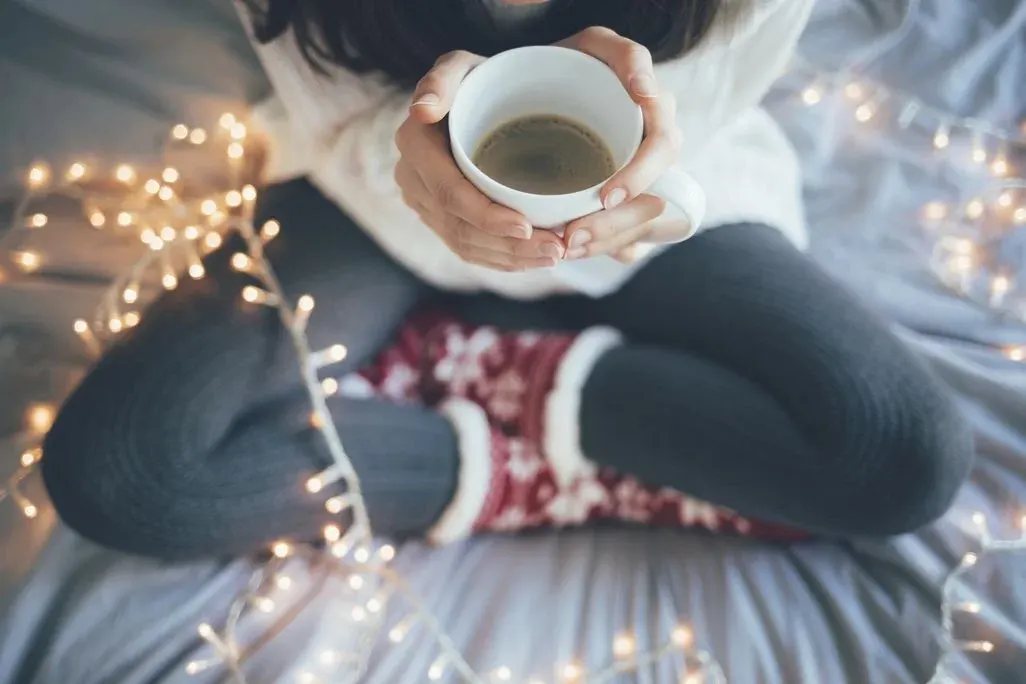 Person in weißem Pullover und grauen Leggings hält weiße Kaffeetasse. Rote Socken und funkelnde Lichterketten umgeben sie.