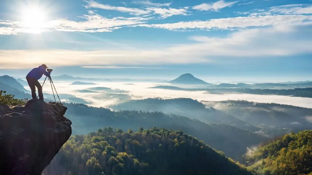 Eine Person mit Kamera auf Stativ steht auf einem Felsen und blickt über nebelverhangene grüne Täler und Berge unter blauem Himmel mit Sonne.