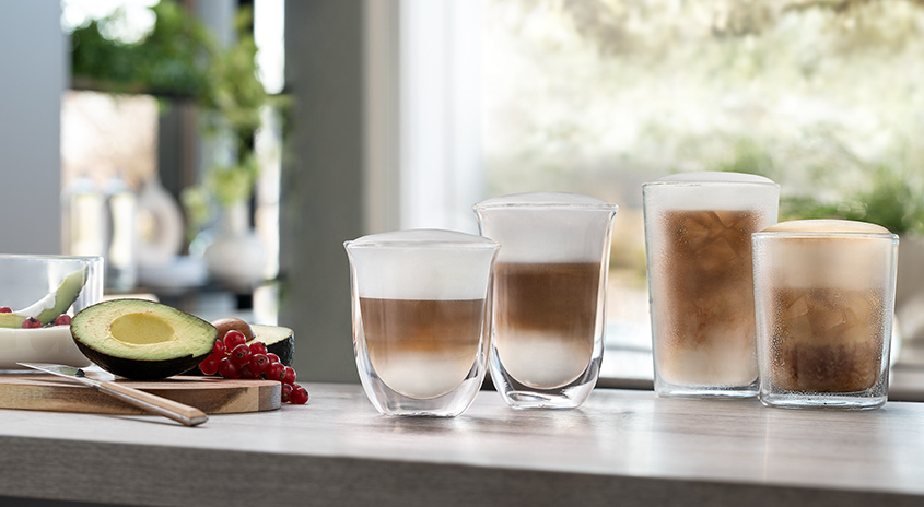 Aufnahme von vier Latte Macchiato-Gläsern, Avocado, Johannisbeeren und einem Messer auf einem Tisch.