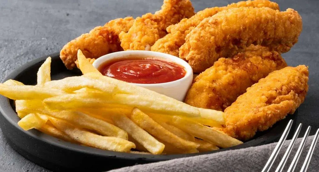 Ein schwarzer runder Teller mit knusprig panierten Chicken Tenders und goldgelben Pommes Frites, serviert mit einem kleinen weißen Schälchen Ketchup in der Mitte. Im Vordergrund rechts liegt eine Serviette mit einer Gabel auf einem grauen Untergrund.