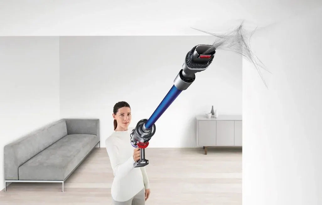 Frau saugt mit kabellosem Staubsauger Spinnweben an der hohen Zimmerdecke über dem Wohnzimmersofa ab.