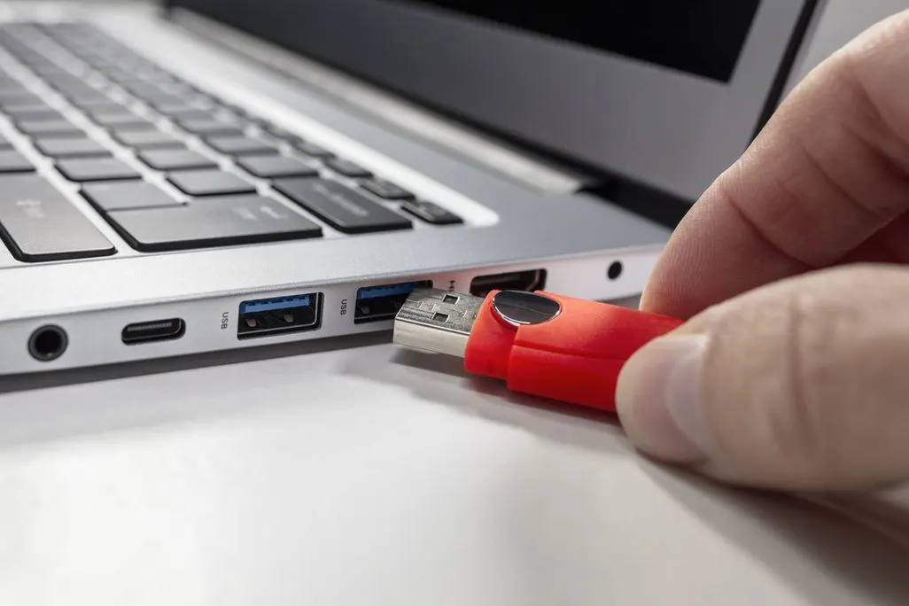 Hand steckt roten USB-Stick in silbernen Laptop mit schwarzer Tastatur. USB-Anschlüsse und Audiobuchse sichtbar.