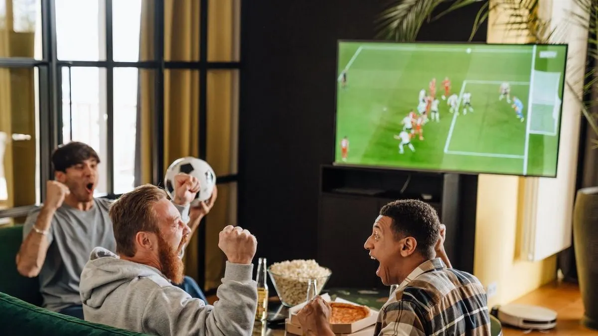 Drei Männer sitzen auf einem Sofa und schauen gemeinsam ein Fußballspiel im Fernsehen mit Snacks und Getränken auf dem Tisch