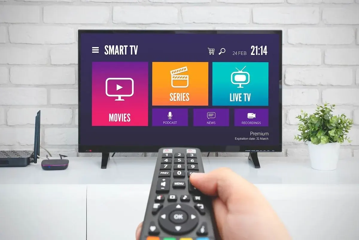 Hand hält Fernbedienung vor Smart TV mit Streaming-Menü. Bildschirm zeigt farbige Kacheln: Movies, Series, Live TV, Podcast, News, Recordings. Wohnzimmer.