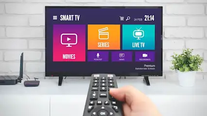 Hand hält Fernbedienung vor Smart TV mit Streaming-Menü. Bildschirm zeigt farbige Kacheln: Movies, Series, Live TV, Podcast, News, Recordings. Wohnzimmer.