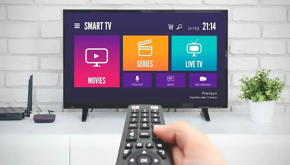 Hand hält Fernbedienung vor Smart TV mit Streaming-Menü. Bildschirm zeigt farbige Kacheln: Movies, Series, Live TV, Podcast, News, Recordings. Wohnzimmer.
