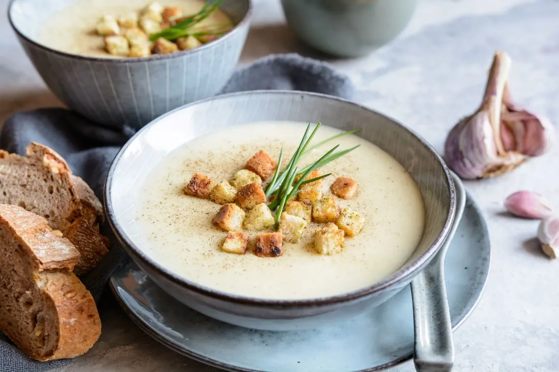 Knoblauchcremesuppe in hellgrauen Teller, getoppt mit Croutons und Schnittlauch, daneben liegt Knoblauch und dunkles Brot