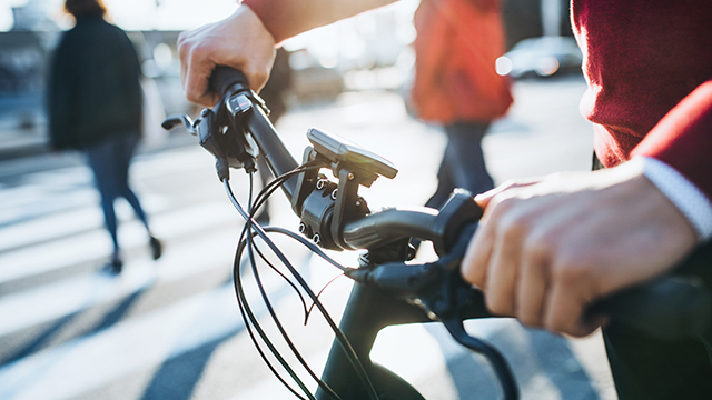 Eine Person hält einen schwarzen Fahrradlenker auf einem Zebrastreifen mit Passanten im Hintergrund.