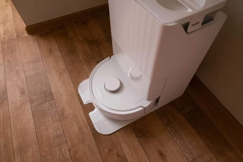 Weißer Saugroboter steht in seiner selbstentleerenden Ladestation auf einem Holzboden im Flur und wird automatisch gereinigt.