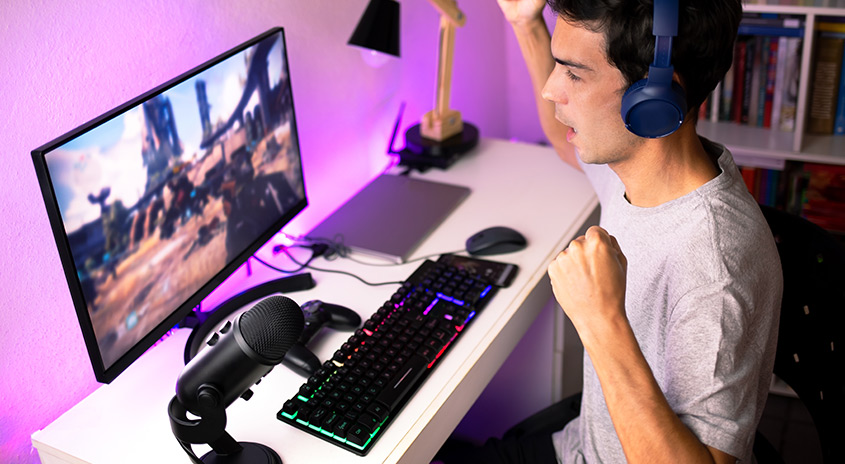 Ein Schreibtisch mit einem Monitor, einer Tastatur, einem Mikrofon und einem Laptop unter violetter Beleuchtung. Ein Regal ist im Hintergrund sichtbar.