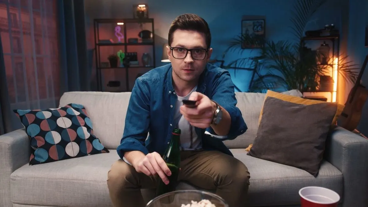 Person sitzt auf einem Sofa und schaut mit Fernbedienung und Getränk in der Hand fern.