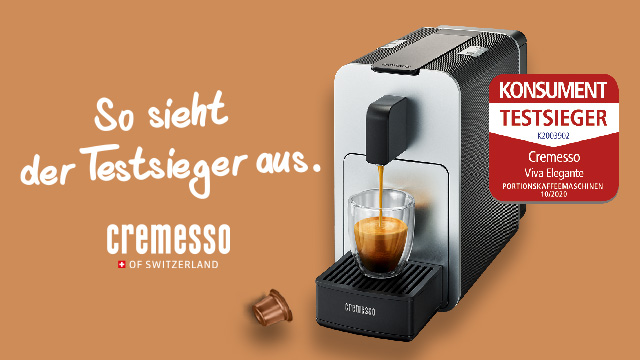 Kaffeemaschine füllt Tasse mit Kaffee. Eine Kapsel liegt daneben. Roter Aufkleber "Testsieger" an der Maschine. Brauner Hintergrund.