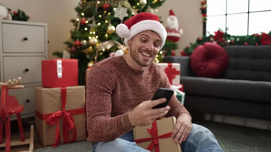 Ein Mann mit roter Weihnachtsmütze und Pullover lächelt, blickt auf ein Telefon und hält ein Geschenk. Im Hintergrund sind ein beleuchteter Weihnachtsbaum, eingepackte Geschenke und ein Sofa.