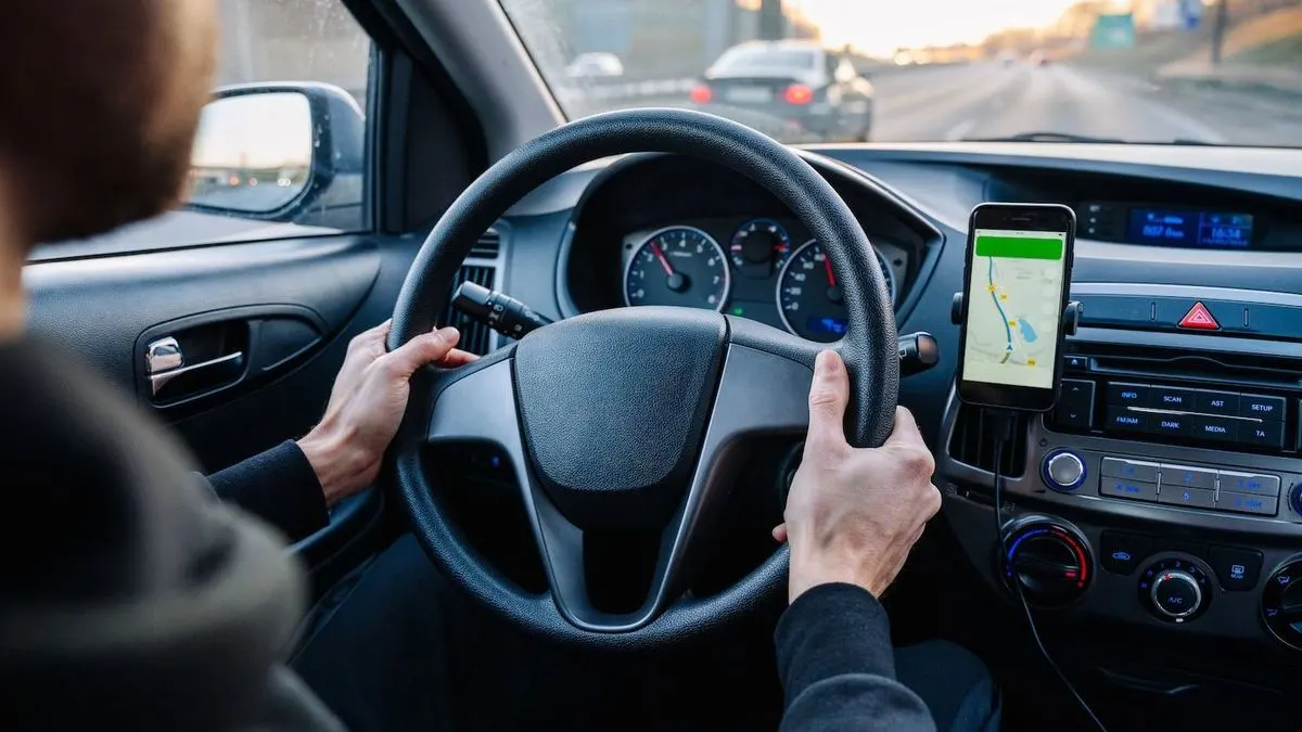 Hände am schwarzen Lenkrad eines Autos. Ein Smartphone mit Navigations-App ist am Armaturenbrett befestigt, Autobahn mit Autos im Hintergrund.