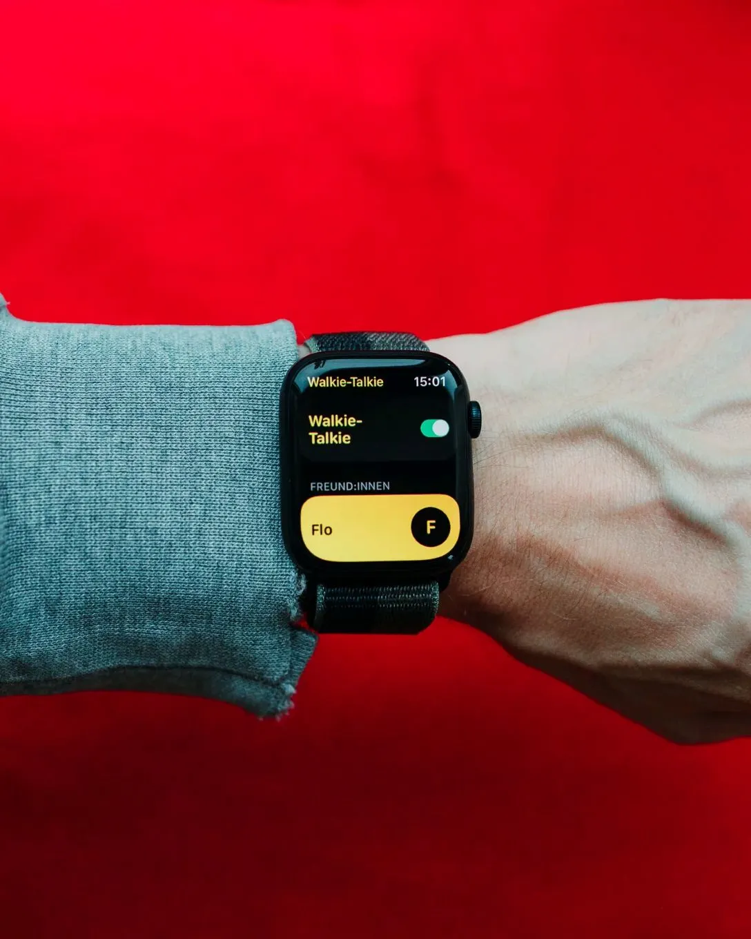 Eine Smartwatch am Handgelenk zeigt die Walkie-Talkie-App mit Kontakt 'Flo' und Uhrzeit 15:01, vor rotem Hintergrund.