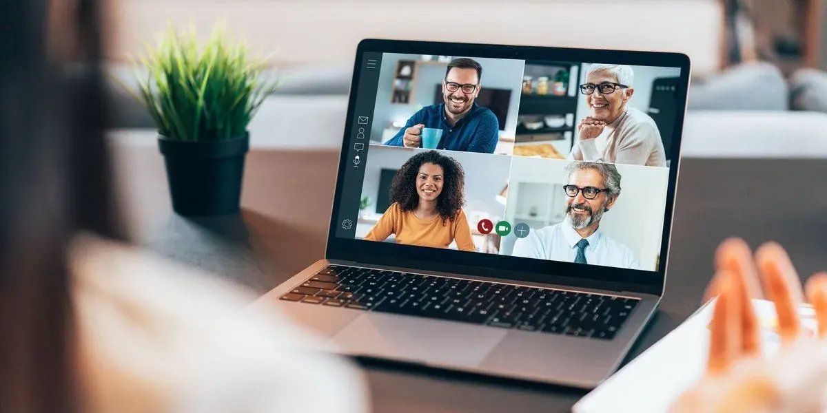 Ein Laptop auf einem dunklen Tisch zeigt eine Videokonferenz mit vier lächelnden Personen. Links steht eine grüne Topfpflanze, rechts sind Hände und ein Dokument.