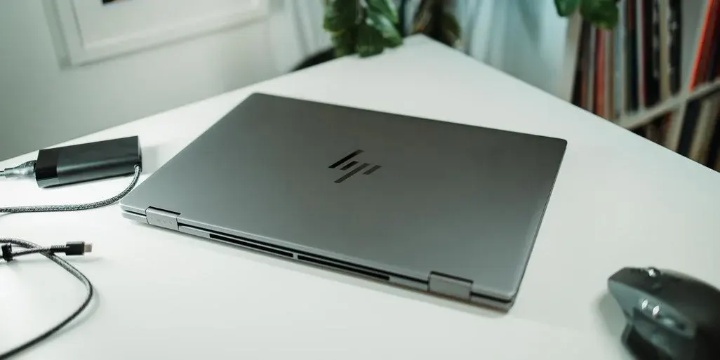 Grauer, geschlossener Laptop mit schwarzem Logo, externer Festplatte und schwarzer Maus auf weißem Tisch.