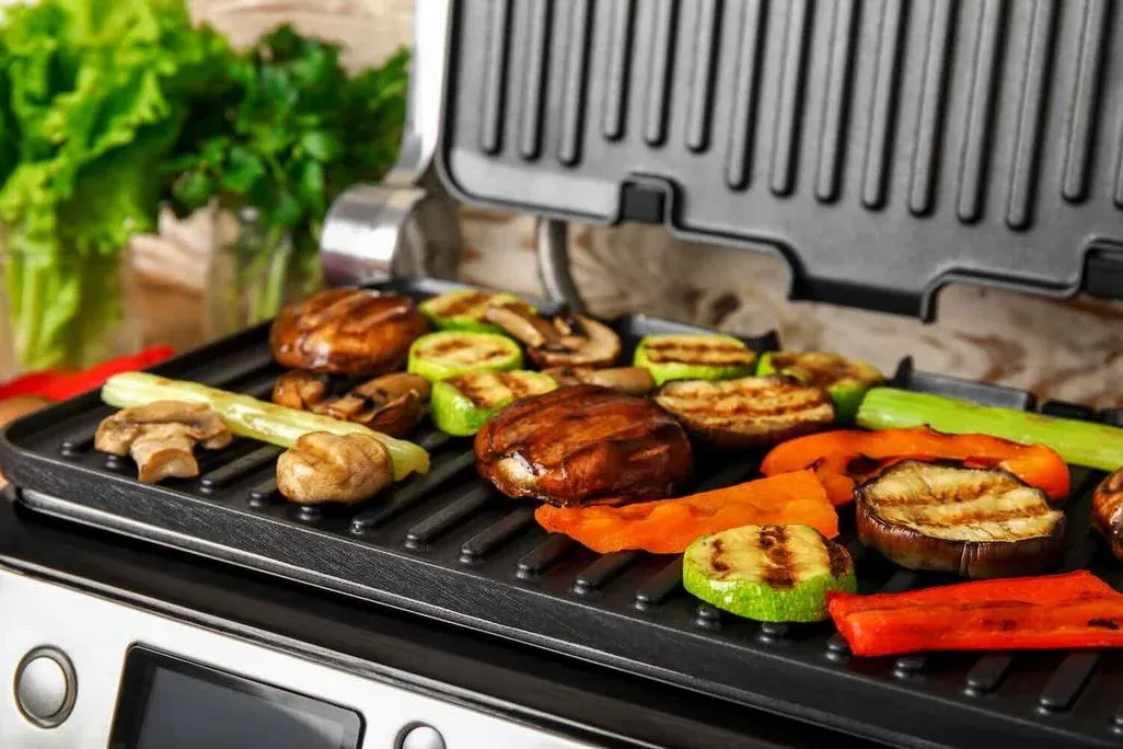 Gegrilltes Gemüse auf einem elektrischen Tischgrill aus Edelstahl mit aufgeklapptem Deckel. Auf der gerillten schwarzen Grillplatte liegen braune Champignons, Zucchinischeiben, rote Paprikastreifen, Auberginenscheiben und Stangensellerie mit sichtbaren Grillmarkierungen. Im Hintergrund sind frischer grüner Salat und Petersilie zu erkennen.