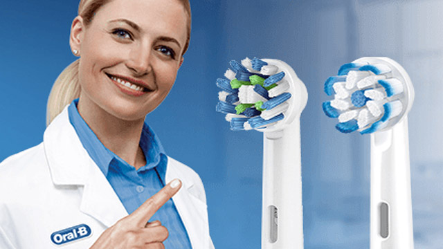 Eine Frau im Laborkittel zeigt auf zwei elektrische Zahnbürstenaufsätze vor blauem Hintergrund.