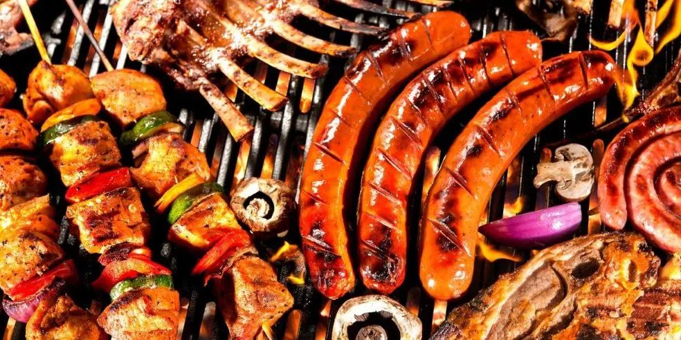 Verschiedenes Grillgut auf einem Rost: braune Würste, Fleischspieße mit Paprika, Rippchen, Pilze und Zwiebeln. Flammen sichtbar.
