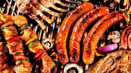 Verschiedenes Grillgut auf einem Rost: braune Würste, Fleischspieße mit Paprika, Rippchen, Pilze und Zwiebeln. Flammen sichtbar.